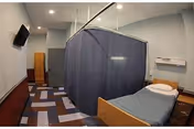 A small room with a single bed featuring a pillow and blanket, a privacy curtain surrounding the bed, a wall-mounted television, and a door at the far end. The floor has a patterned carpet and the walls are painted light blue.