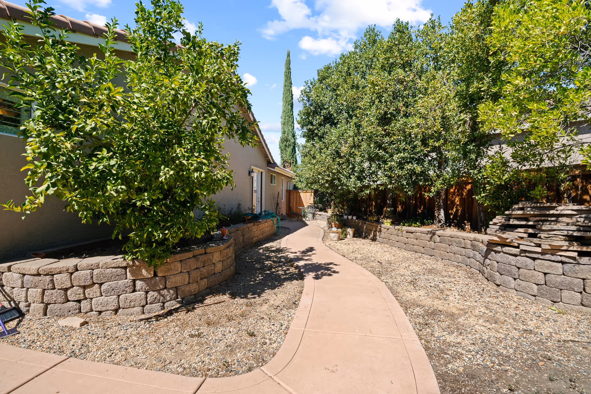 A curved concrete pathway runs between two raised garden beds made of stone blocks. The garden beds contain green leafy trees and plants. A beige building with a tiled roof is on the left side, and a wooden fence is visible on the right side. The sky is blue with some clouds.