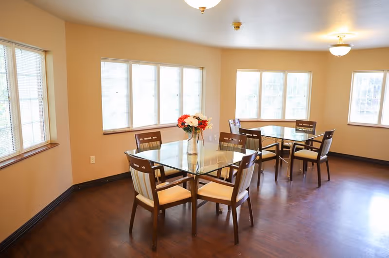 A bright dining room with two glass-top tables, each surrounded by four wooden chairs with beige cushions. A vase with red and white flowers is placed on the table in the foreground. The room has large windows with blinds, beige walls, and dark wooden flooring.