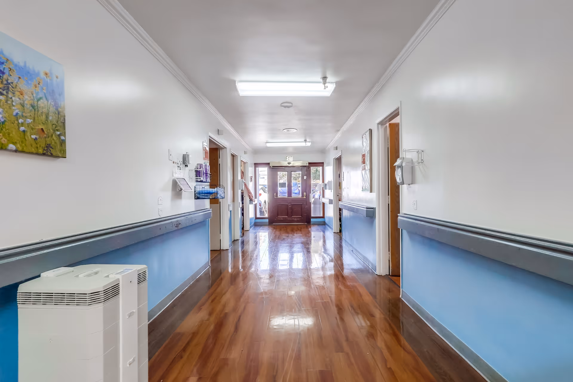 A clean and well-lit hallway in a senior living facility with wooden flooring, blue and white walls, handrails on both sides, and several open doorways leading to rooms. There is a painting of flowers on the left wall and an air purifier on the floor. The hallway ends with a glass door letting in natural light.
