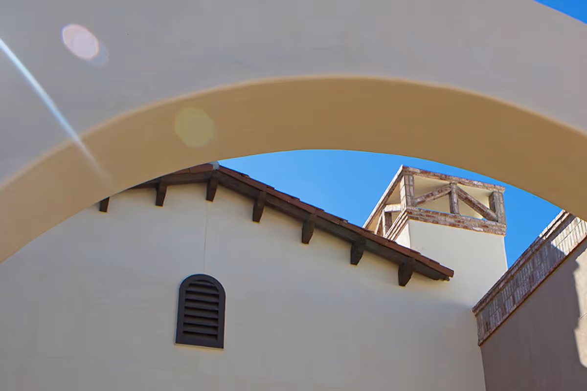 Stucco building rooflines and a small tower seen through a large arch against a blue sky.