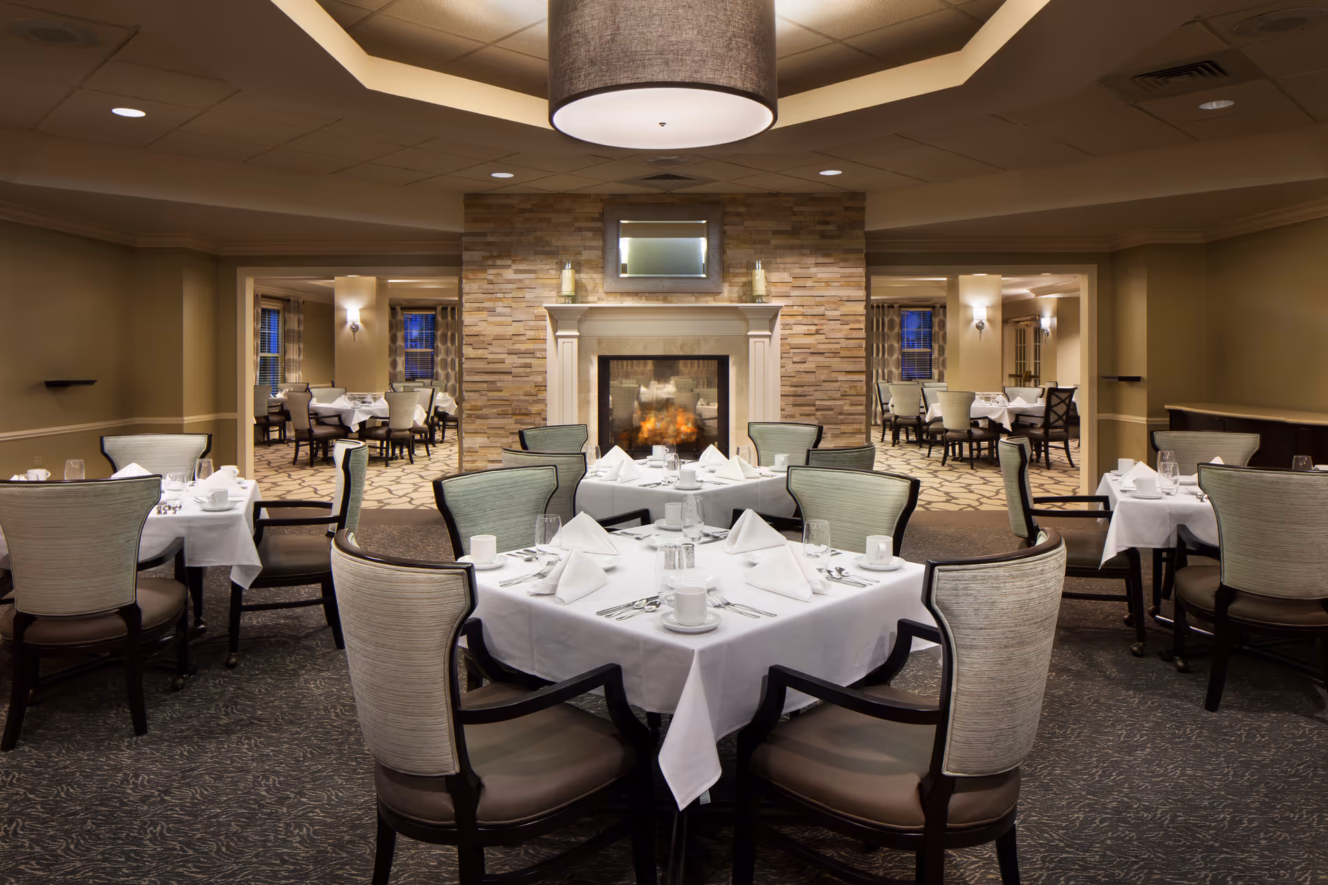A warmly lit dining room with tables covered in white tablecloths, neatly set with napkins, cups, glasses, and silverware. The room features a stone fireplace with a fire burning and a mirror above it. Additional dining tables and chairs are visible through open doorways on either side of the fireplace.
