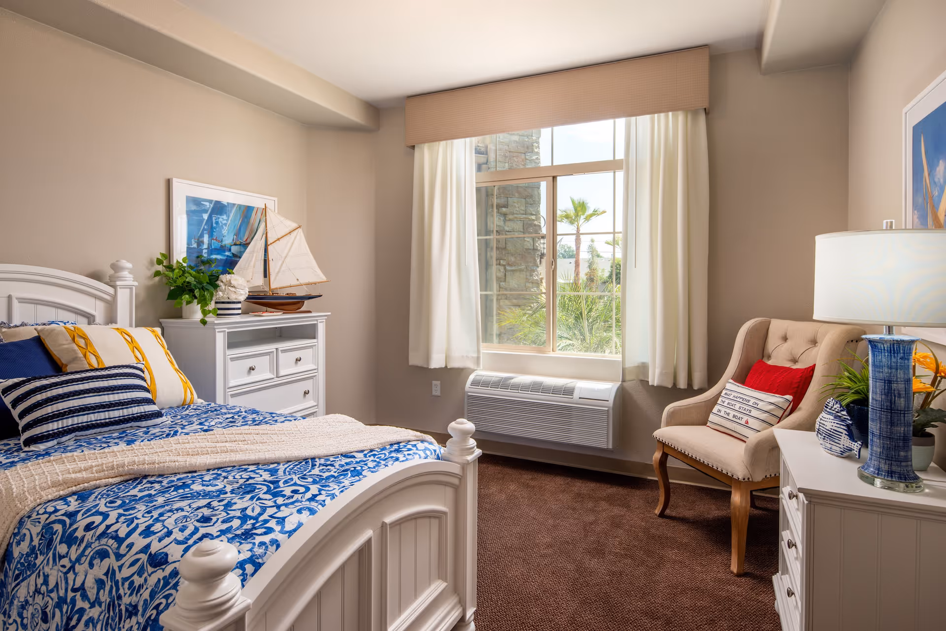 A cozy bedroom with a white wooden bed featuring blue and white patterned bedding and multiple pillows. There is a white dresser with a decorative sailboat and a plant on top. A window with white curtains lets in natural light, showing palm trees outside. A beige armchair with a red pillow and a pillow with text sits next to a white nightstand with a blue lamp and more decorative items.