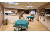 Interior view of a senior living facility dining area with light wood flooring and beige walls. There are two round tables covered with green tablecloths, each surrounded by metal chairs. The room includes a kitchenette with cabinets, a countertop, a microwave, and a refrigerator. The ceiling has recessed lighting and a drop ceiling design.