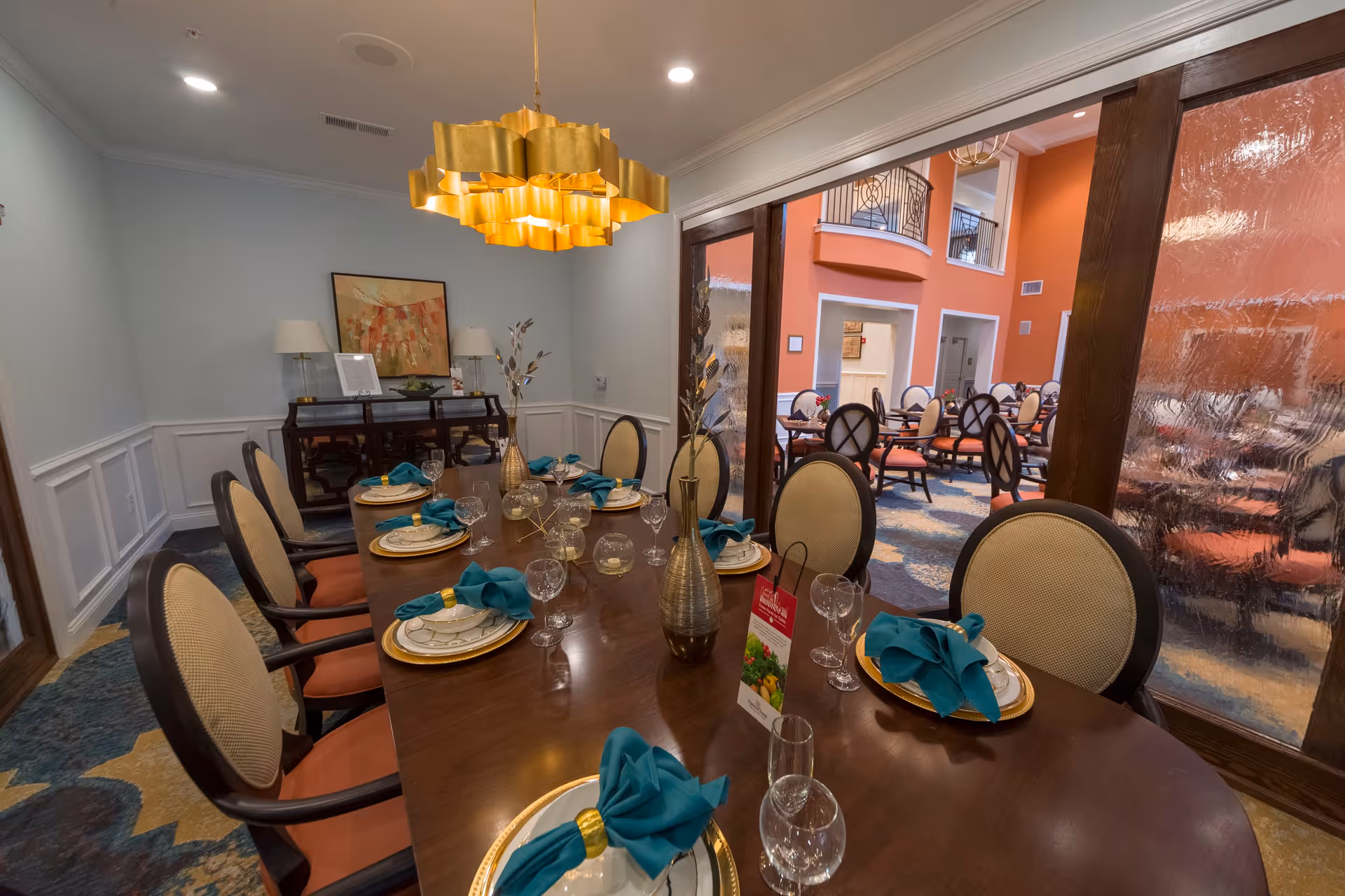 A formal dining room with a long wooden table set for eight people with plates, glasses, and teal napkins. The room features a gold chandelier, a sideboard with lamps and artwork, and a view into a larger dining area with multiple tables and chairs through glass panels.