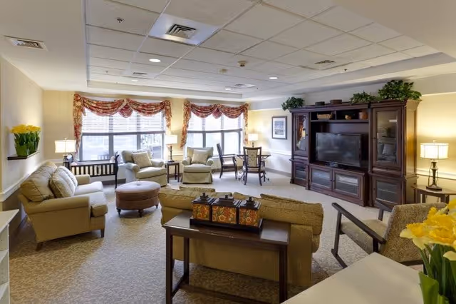 A well-lit living room in a senior living facility with beige sofas and armchairs arranged around a central ottoman. There is a large wooden entertainment center with a flat-screen TV, several lamps providing warm lighting, and windows with red and gold valances. The room is decorated with plants and framed artwork, creating a cozy and inviting atmosphere.