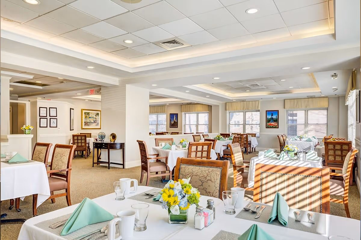 Bright, well-lit dining room with neatly set tables, floral centerpieces, and upholstered chairs.