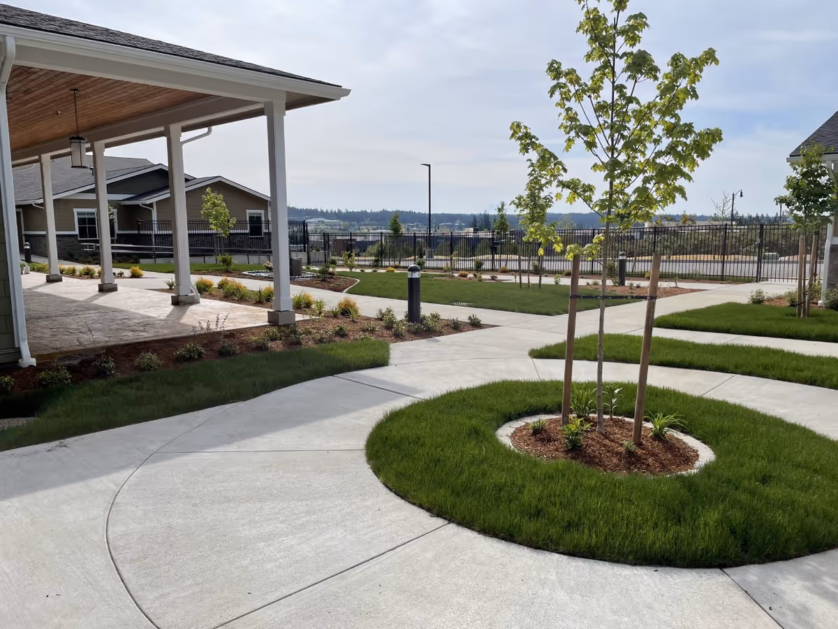 Outdoor area of a senior living facility with concrete walkways curving around small landscaped grassy areas with young trees. There is a covered patio with wooden ceiling and hanging lantern-style lights. In the background, there are residential-style buildings and a black metal fence with a view of distant hills under a partly cloudy sky.