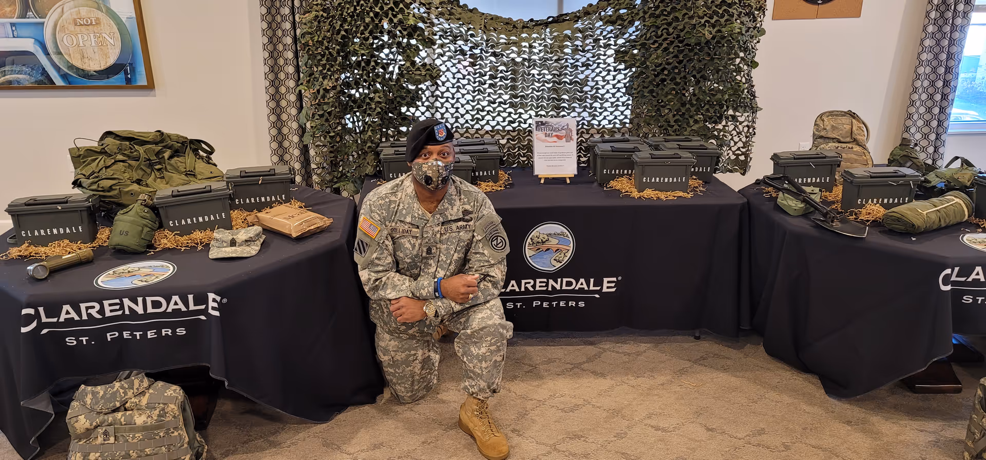 A person dressed in a U.S. Army uniform kneels on the floor in front of three tables covered with black tablecloths labeled 'Clarendale St. Peters'. The tables display military-themed items including green ammunition boxes, backpacks, helmets, and other gear. Behind the tables is a camouflage net hanging on the wall.