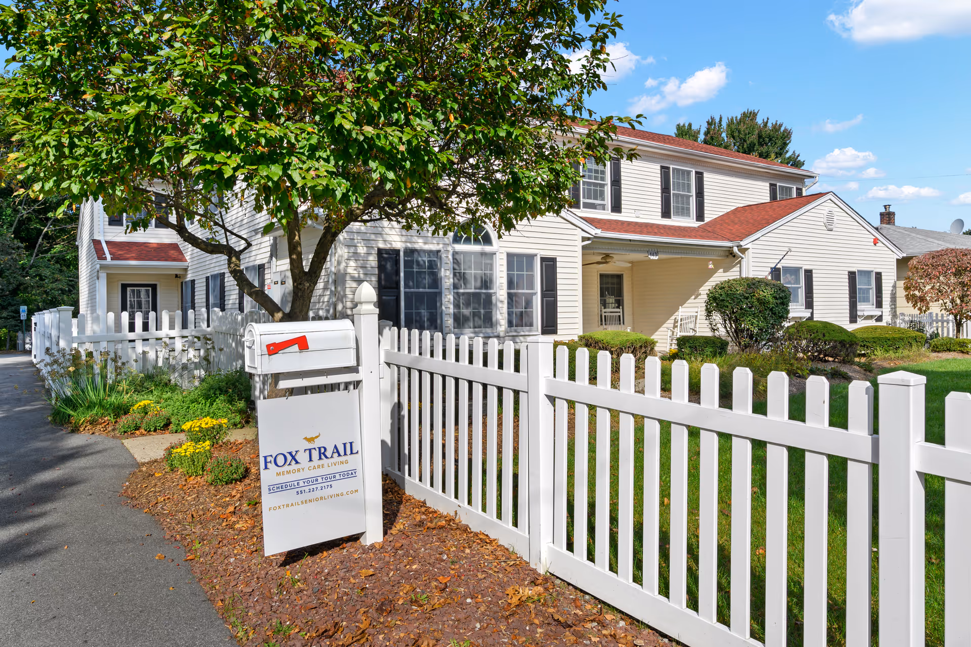 Exterior view of Fox Trail Memory Care at Paramus, showing a white two-story building with black shutters and a red roof. A white picket fence runs along the front yard with green grass and bushes. A white mailbox and a sign with the facility's name and contact information are visible near the sidewalk. A tree with green leaves partially shades the area.