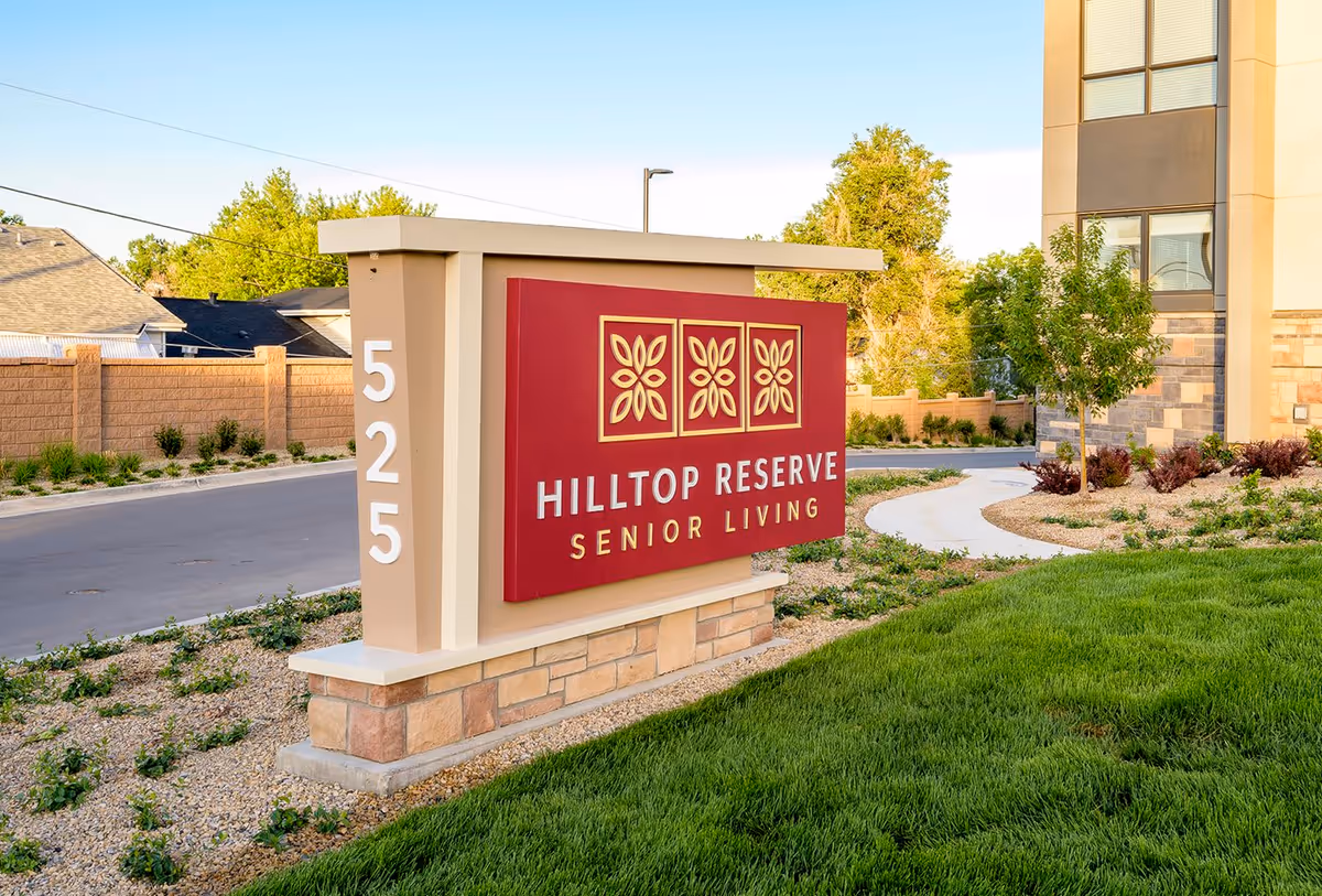 Outdoor view of a sign for Hilltop Reserve Senior Living with the address number 525 displayed on a beige and stone base structure, surrounded by landscaped greenery and a building partially visible in the background.