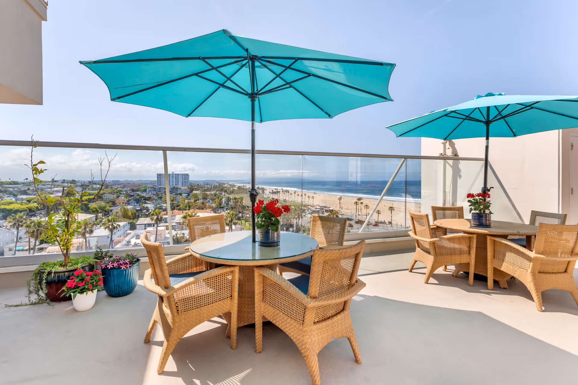 Outdoor patio area with two round glass-top tables, each surrounded by four wicker chairs. Each table has a turquoise umbrella providing shade and a small potted plant with red flowers in the center. The patio overlooks a scenic view of a beach, ocean, and nearby buildings under a clear blue sky.