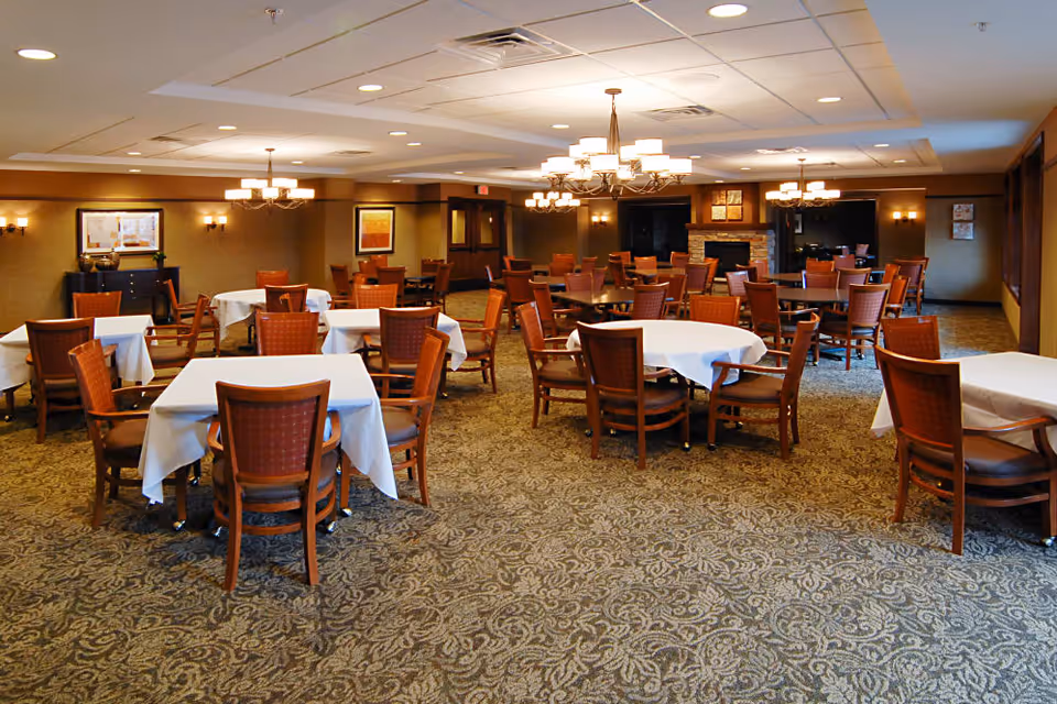 A spacious dining room with multiple tables covered in white tablecloths and surrounded by wooden chairs with red cushions. The room features patterned carpet flooring, warm lighting from chandeliers and wall sconces, and framed artwork on the walls. There is a fireplace in the background and large windows on the right side.