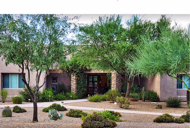 Outdoor view of a senior living facility with desert landscaping including various shrubs, cacti, and trees. The building has a beige exterior with a stone pillar entrance and windows on either side.