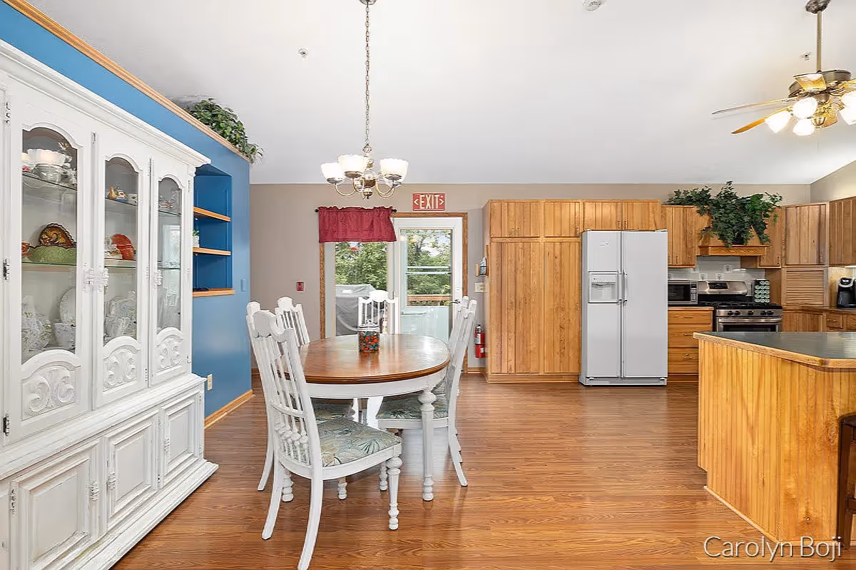 A bright kitchen and dining area with wooden floors and cabinetry. The dining area features a wooden table with white chairs, a white china cabinet with glass doors, and a chandelier overhead. The kitchen has a white refrigerator, stove, microwave, and a ceiling fan with lights. There is a door with a red curtain leading outside, and some greenery decorates the top of the cabinets.