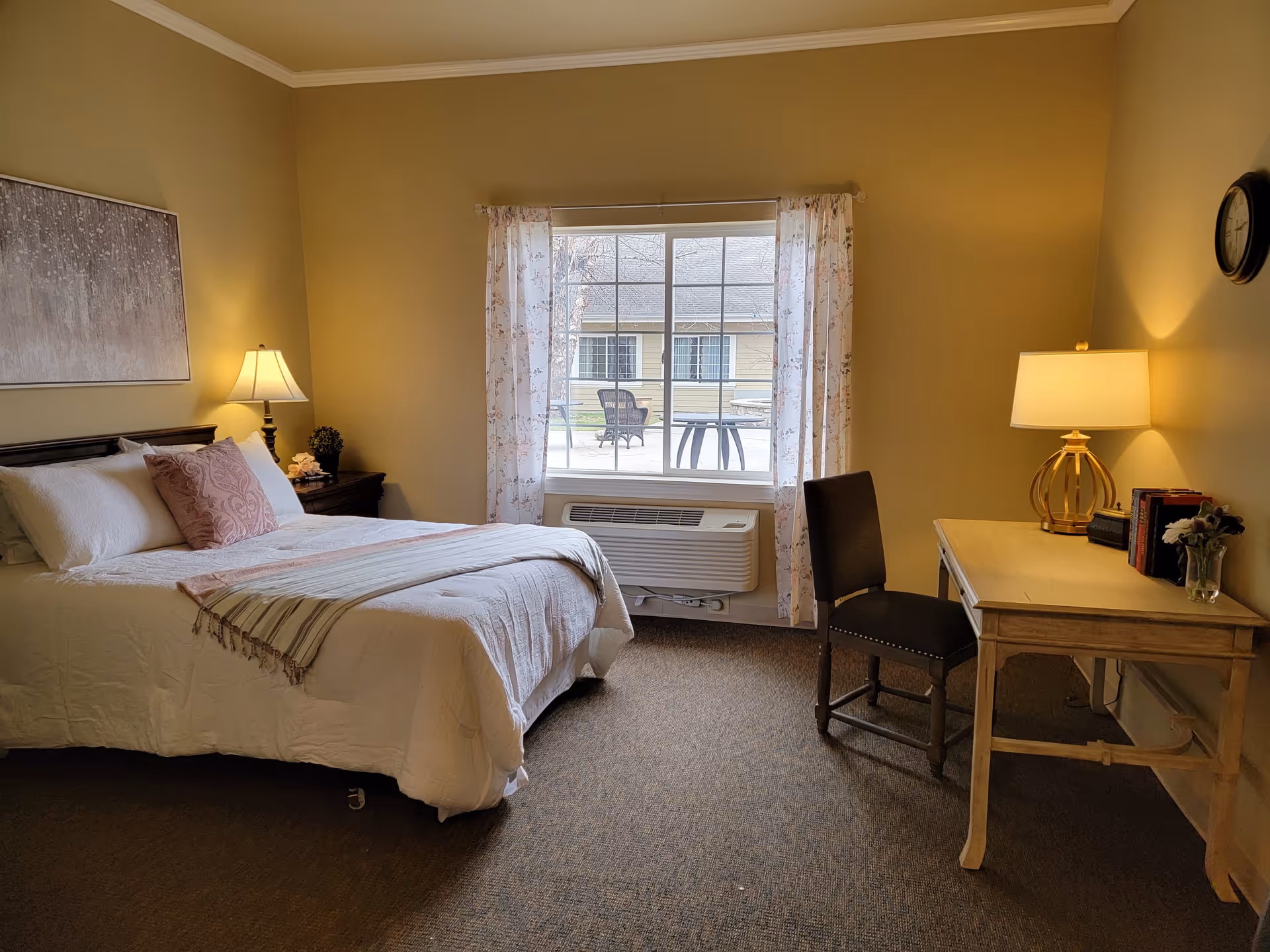 A cozy bedroom with a neatly made bed featuring white bedding and a pink decorative pillow. There is a wooden nightstand with a lamp and small decorative items next to the bed. A window with sheer floral curtains lets in natural light and shows an outdoor patio area with chairs and a table. On the right side of the room, there is a wooden desk with a lamp, books, and a small vase with flowers, accompanied by a dark upholstered chair. The walls are painted a warm beige color, and a clock hangs on the wall above the desk.