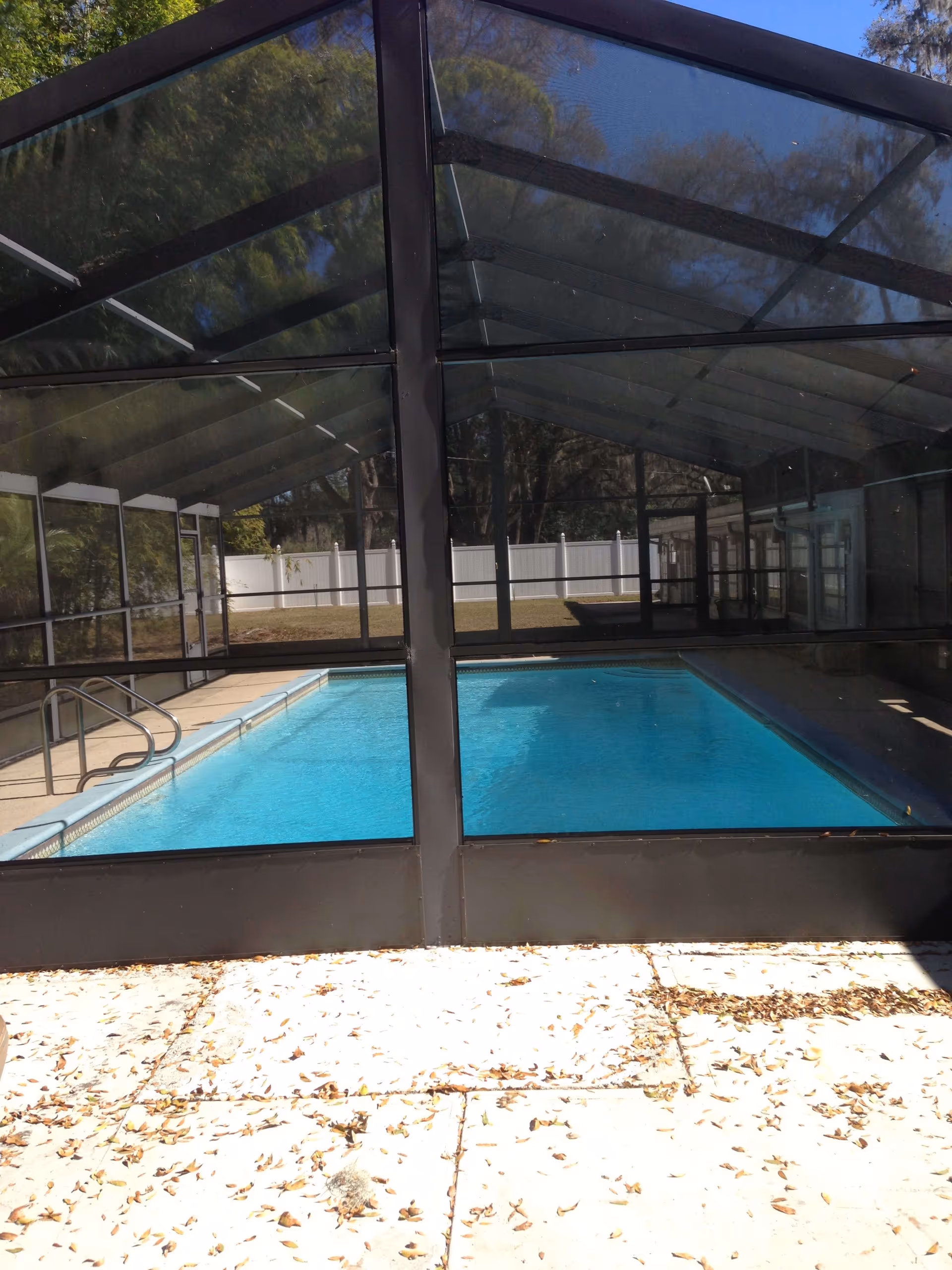 An indoor swimming pool enclosed by a black metal and glass structure. The pool has clear blue water and a metal handrail on the left side. Outside the enclosure, there is a concrete patio with scattered dry leaves and a white fence in the background with trees beyond it.