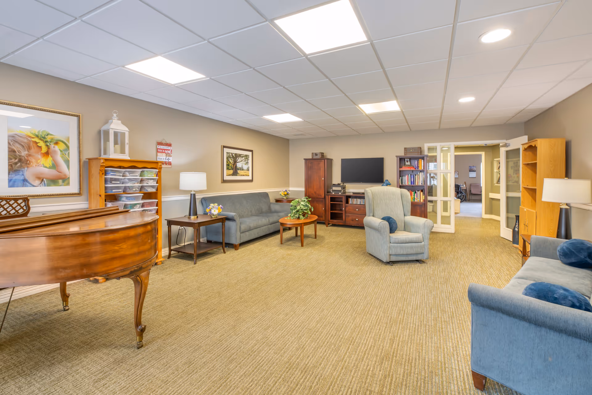 A spacious living room with beige walls and carpeted floor, featuring two blue sofas, a blue armchair, a wooden coffee table with a plant, a wooden side table with a lamp, a wooden piano, a wooden cabinet with a flat-screen TV, and bookshelves. The room is well-lit with ceiling lights and has framed artwork on the walls.