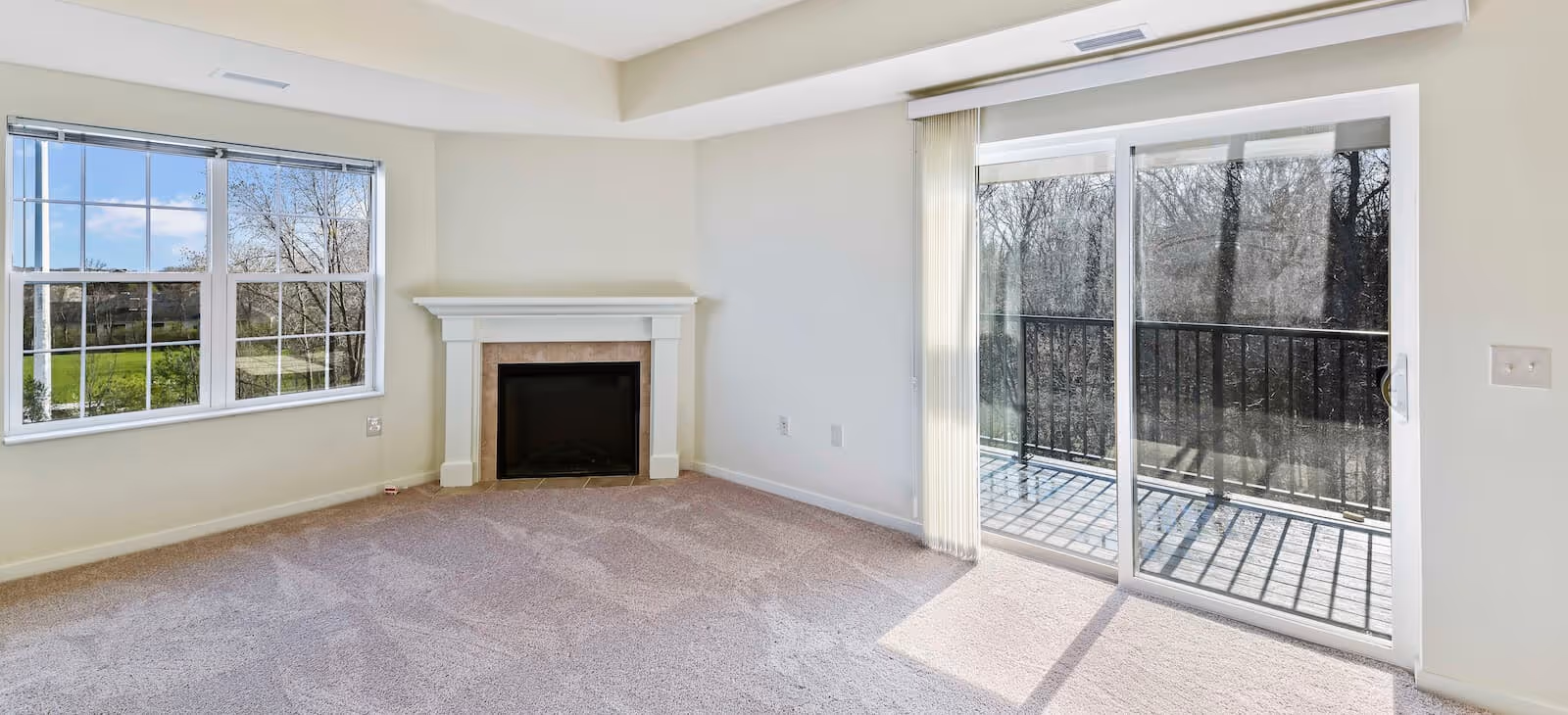 Empty living room with beige carpet, a white fireplace, large window with a view of greenery, and sliding glass doors leading to a balcony overlooking trees.
