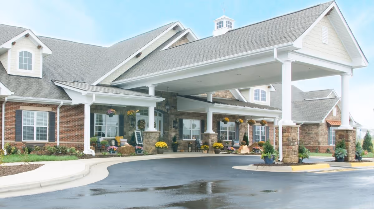 Front exterior view of Bickford of Spotsylvania facility showing a covered entrance with stone pillars, brick and stone walls, windows with shutters, and landscaped greenery and flowers.