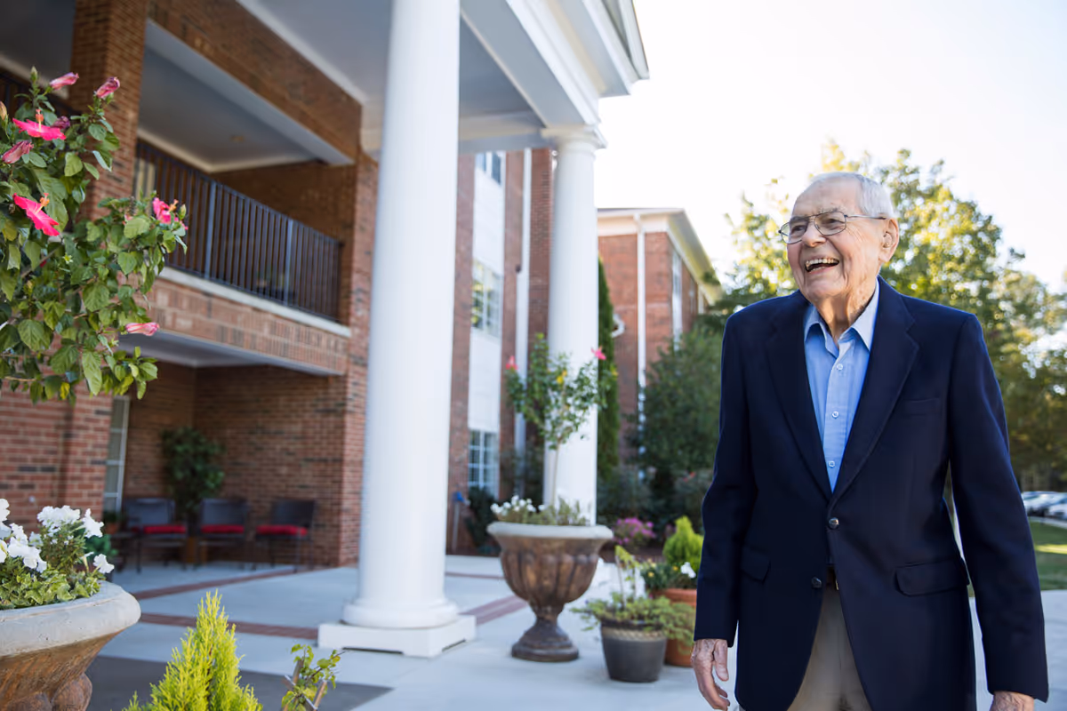 An elderly man wearing a navy blazer and light blue shirt is smiling and standing outside a brick building with white columns and potted plants around the entrance.