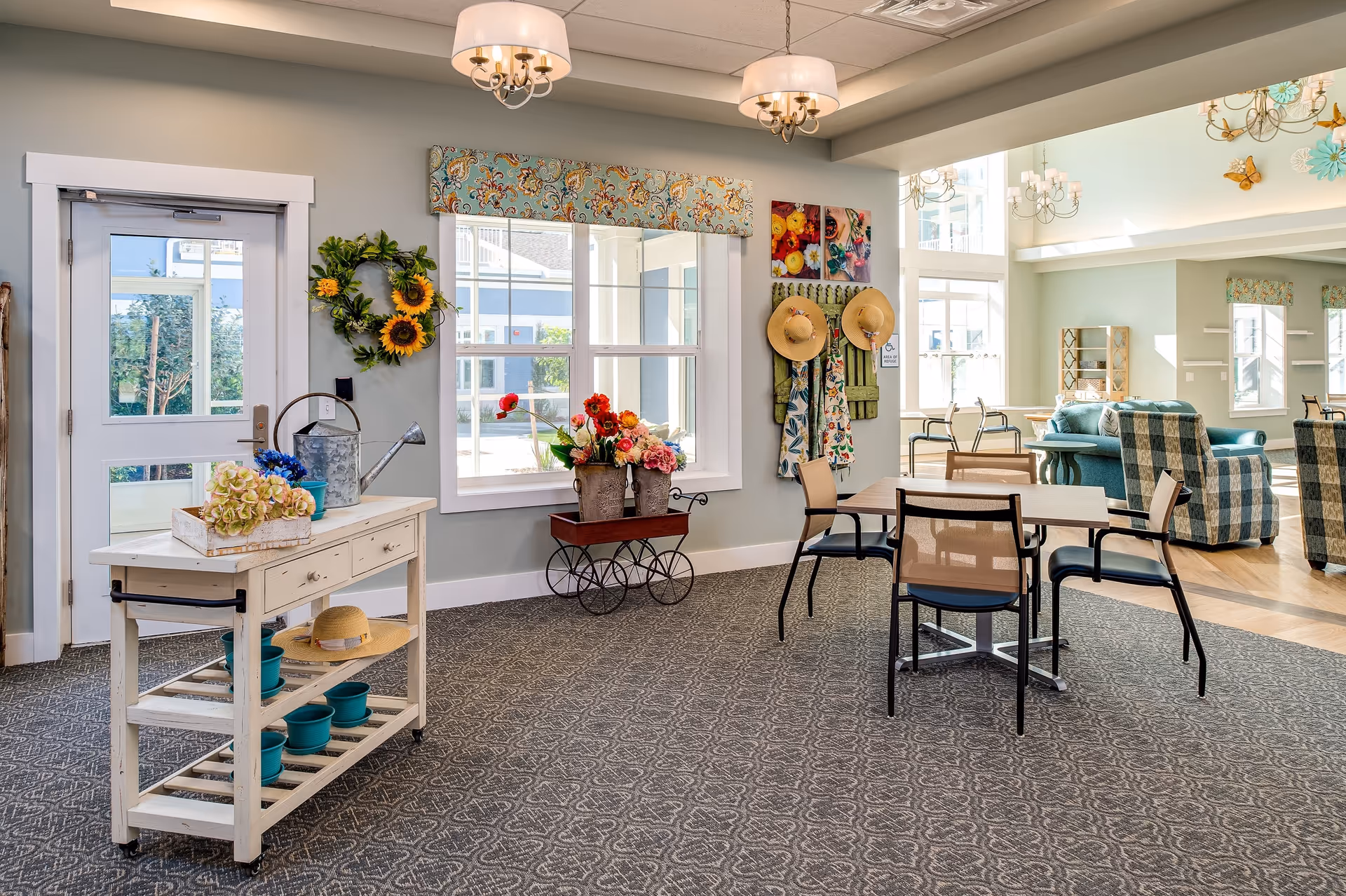 A bright and spacious common area in a senior living facility with a small table and chairs, a white wooden cart with flower pots and a watering can, floral decorations including a sunflower wreath and a cart with colorful flowers, and a cozy seating area with teal and plaid upholstered chairs. Large windows allow natural light to fill the room.