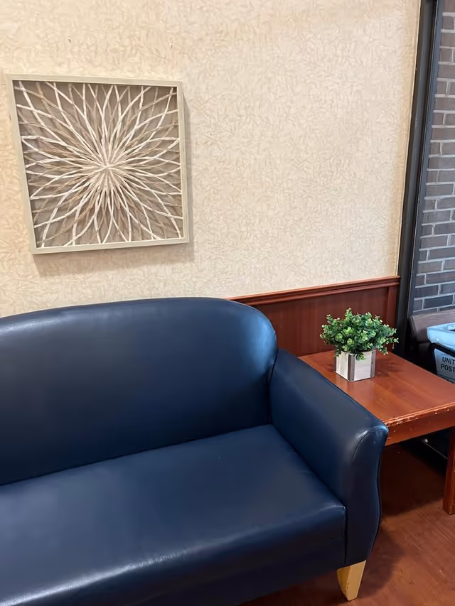 A small seating area with a dark blue leather loveseat, a wooden side table with a small green potted plant, and a decorative wall art piece with a geometric pattern on a beige textured wall. A window with a view of a brick wall is visible on the right side.