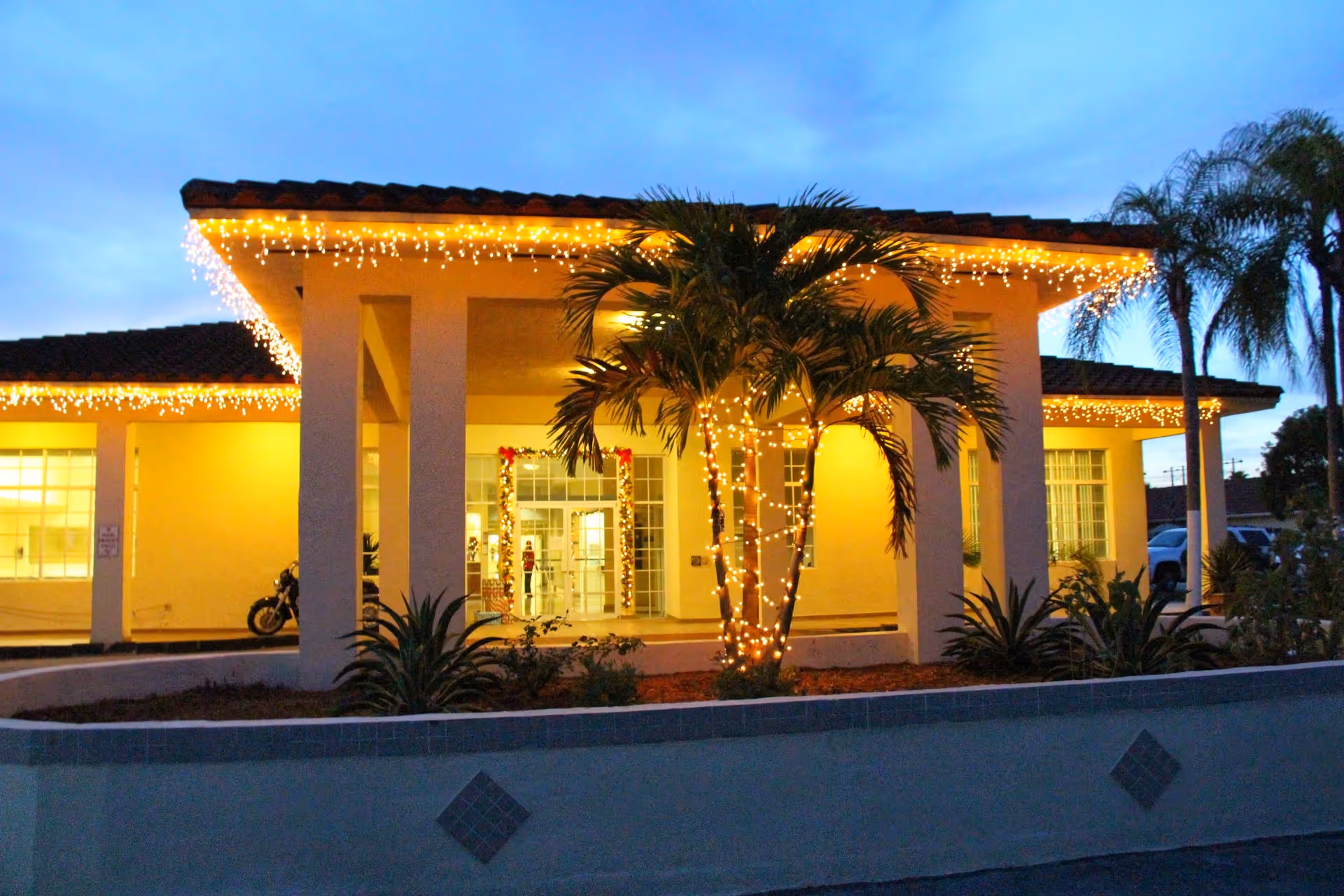 Front entrance of a residential building at dusk decorated with string lights and palm trees.