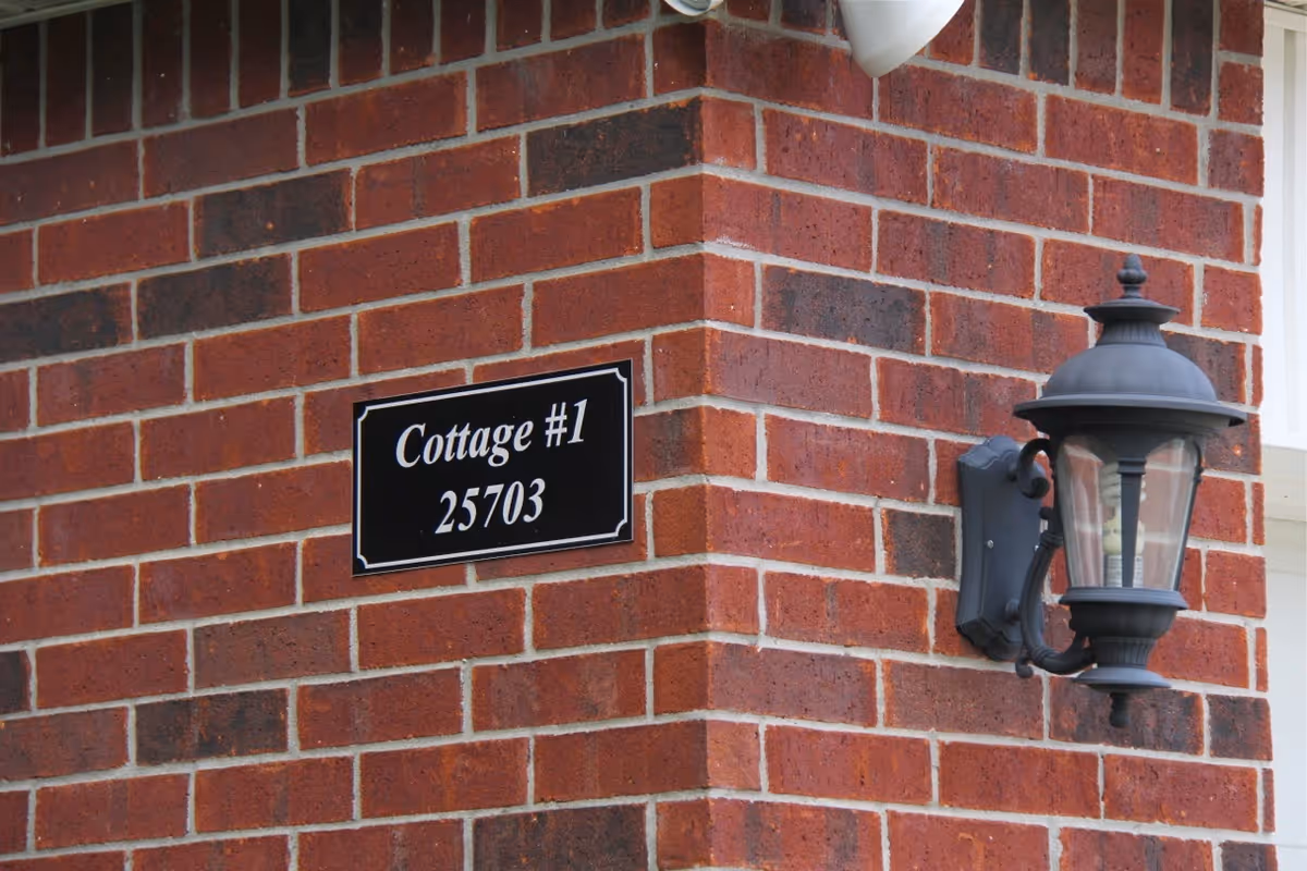 A close-up view of a red brick wall corner with a black and white sign that reads 'Cottage #1 25703' and a black outdoor wall lantern mounted next to the sign.