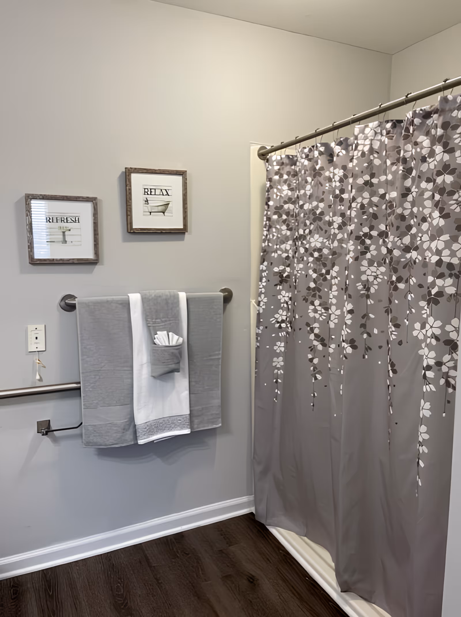 A bathroom corner featuring a shower with a gray floral-patterned curtain, two framed pictures on the wall with the words 'REFRESH' and 'RELAX', a towel rack holding gray and white towels, and a dark wood floor.