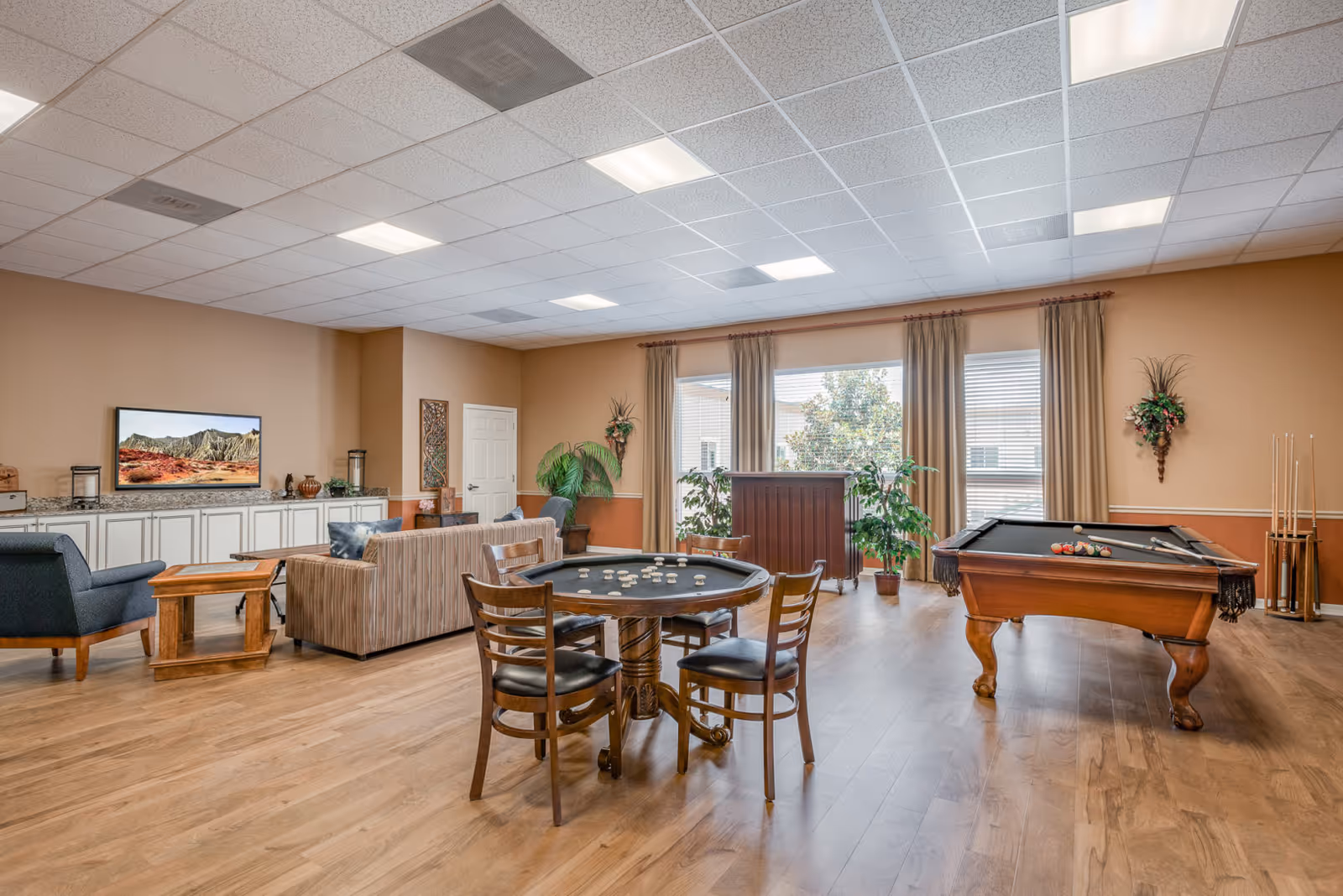 A spacious senior living facility common area with a pool table, a game table with chairs, a striped sofa, armchairs, a large window with beige curtains, and a TV mounted on a beige wall above white cabinets.