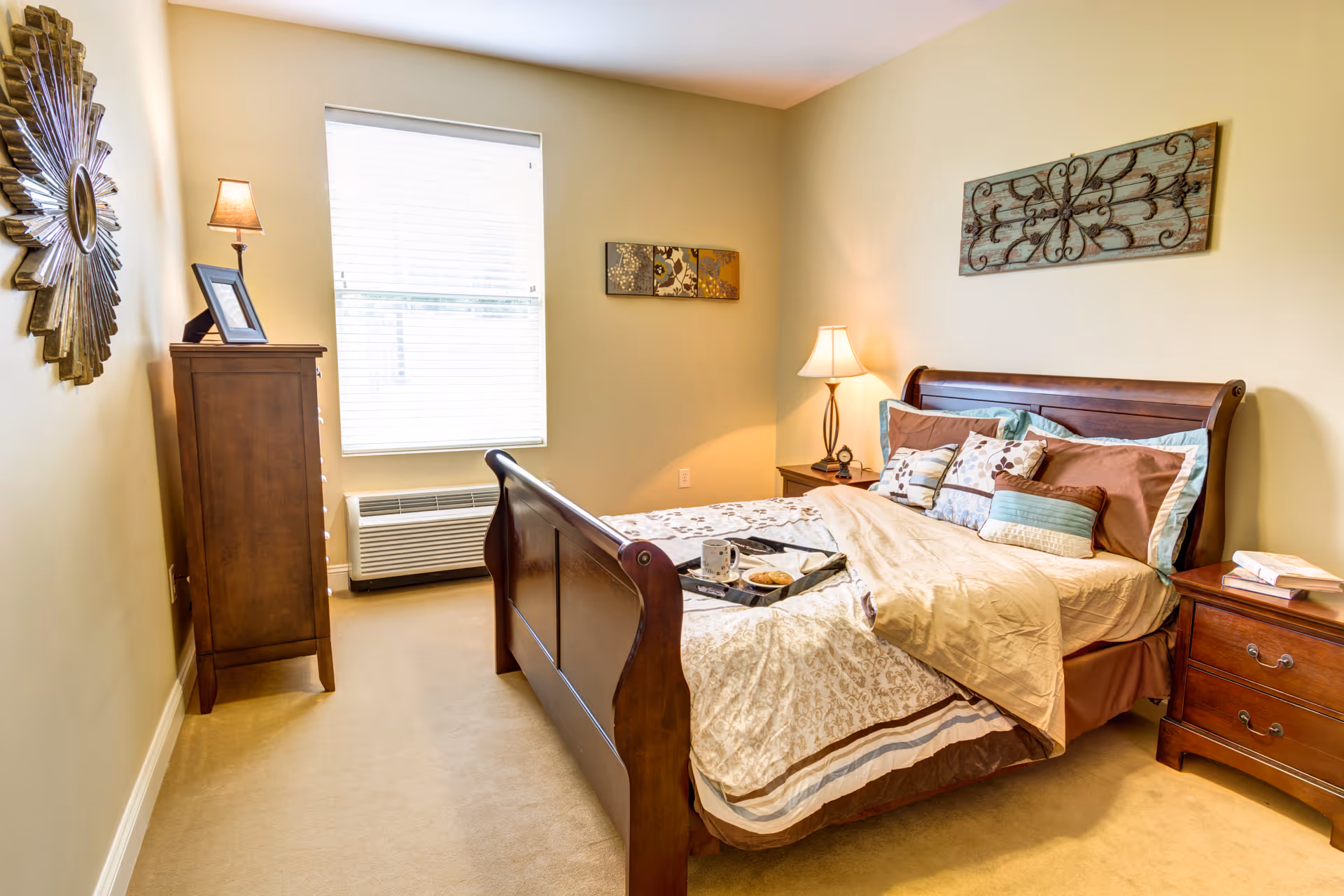 A cozy bedroom with a wooden bed frame, neatly made bed with multiple pillows and a beige comforter. There is a wooden nightstand with a lamp and books on the right side of the bed. A wooden dresser with a small lamp and picture frame is positioned against the left wall. A window with blinds is centered on the back wall, and decorative wall art is hung above the bed and on the left wall. The room has beige walls and carpeted floor.