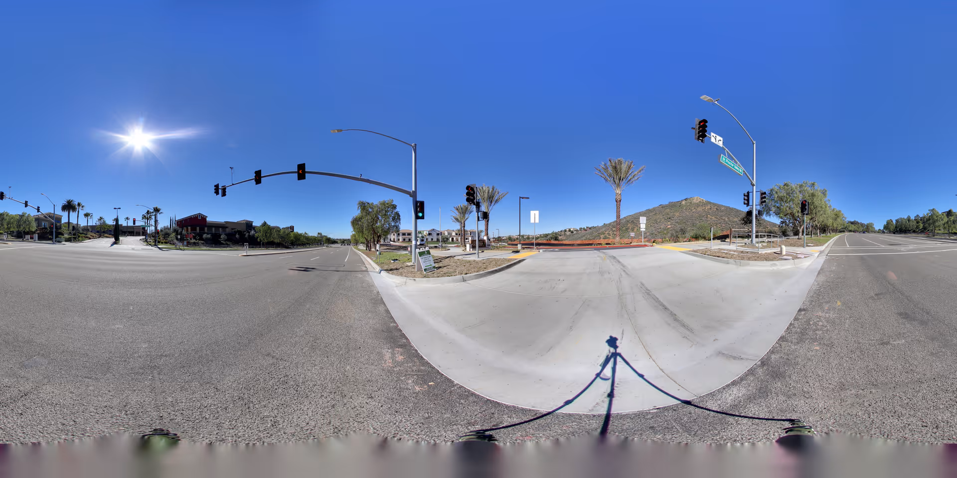 A wide panoramic view of a sunny intersection near Westmont at San Miguel Ranch, featuring traffic lights, palm trees, a clear blue sky, and a hill in the background.