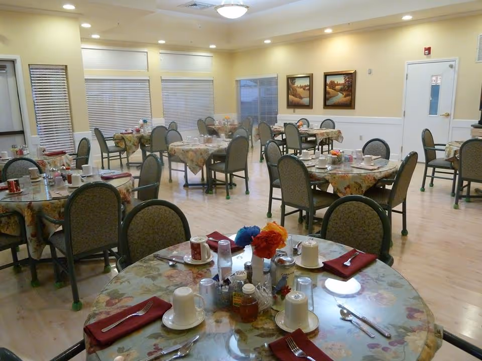 A dining room with multiple round tables covered with floral tablecloths, each set with cups, utensils, napkins, and condiments. The room has light-colored walls, several windows with blinds, framed landscape paintings on the wall, and overhead lighting.