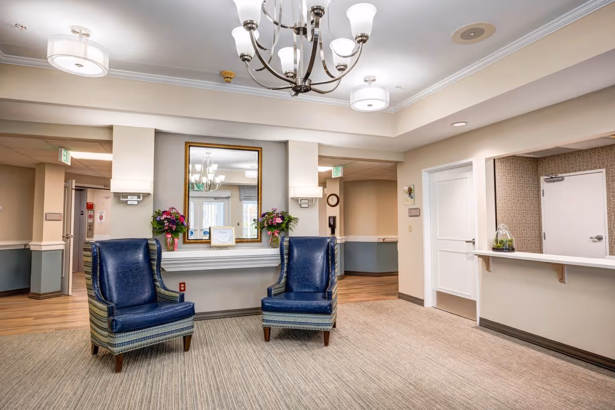 A well-lit interior common area with two blue upholstered armchairs placed on a carpeted floor. Behind the chairs is a wall with a large mirror framed in gold, flanked by two wall sconces and flower arrangements on a narrow shelf. To the right, there is a reception counter with a small plant decoration and a closed white door behind it. The walls are painted in neutral tones with a chair rail and wood flooring visible in the adjacent hallway.