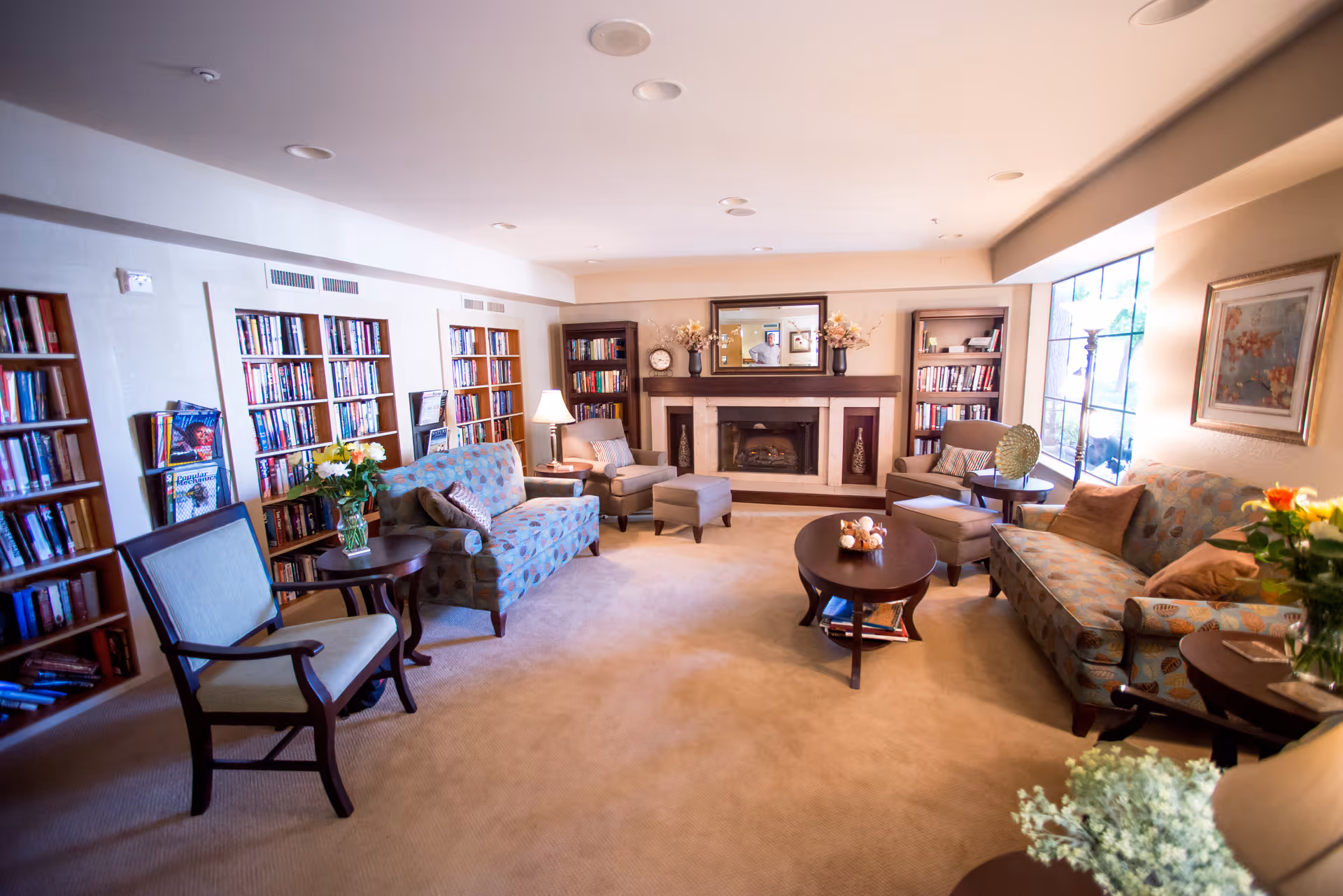 A cozy living room with beige carpet, two patterned sofas, two armchairs with matching ottomans, a wooden coffee table, and several bookshelves filled with books. There is a fireplace with a mirror above it and decorative vases on the mantel. A large window lets in natural light, and there are several lamps and floral arrangements around the room.