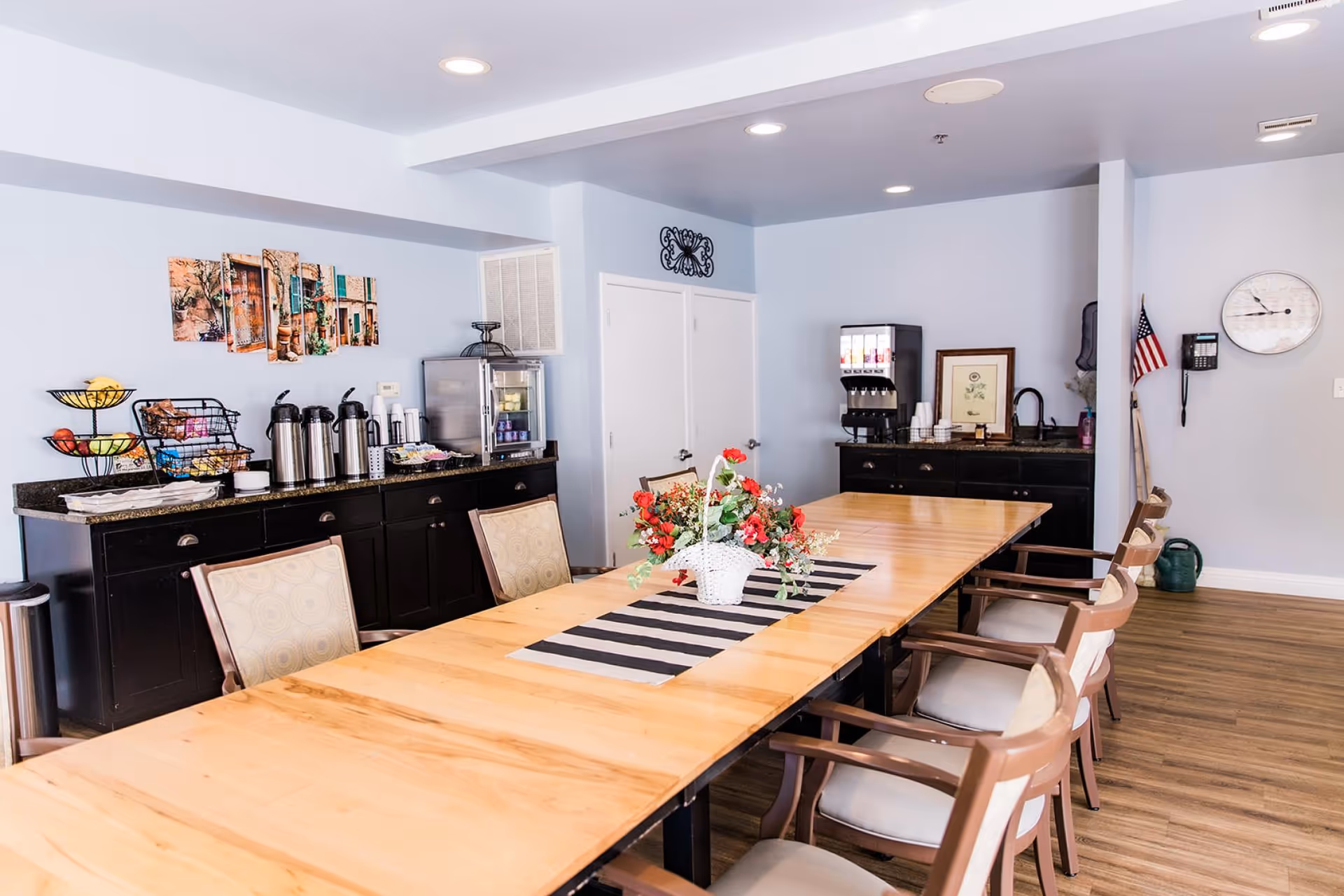 A bright dining room with a long wooden table surrounded by chairs. The table has a black and white striped runner with a white basket filled with red and white flowers as a centerpiece. Along the walls, there are black cabinets with various beverage dispensers, coffee pots, snacks, and cups. The room has light blue walls, a wall clock, an American flag, and a decorative wall piece above double doors.