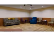 A spacious room with wooden flooring and beige walls featuring a patterned sofa with two yellow pillows, a blue recliner chair, and several wooden chairs arranged along the walls. The ceiling has recessed lighting and a ceiling fan.