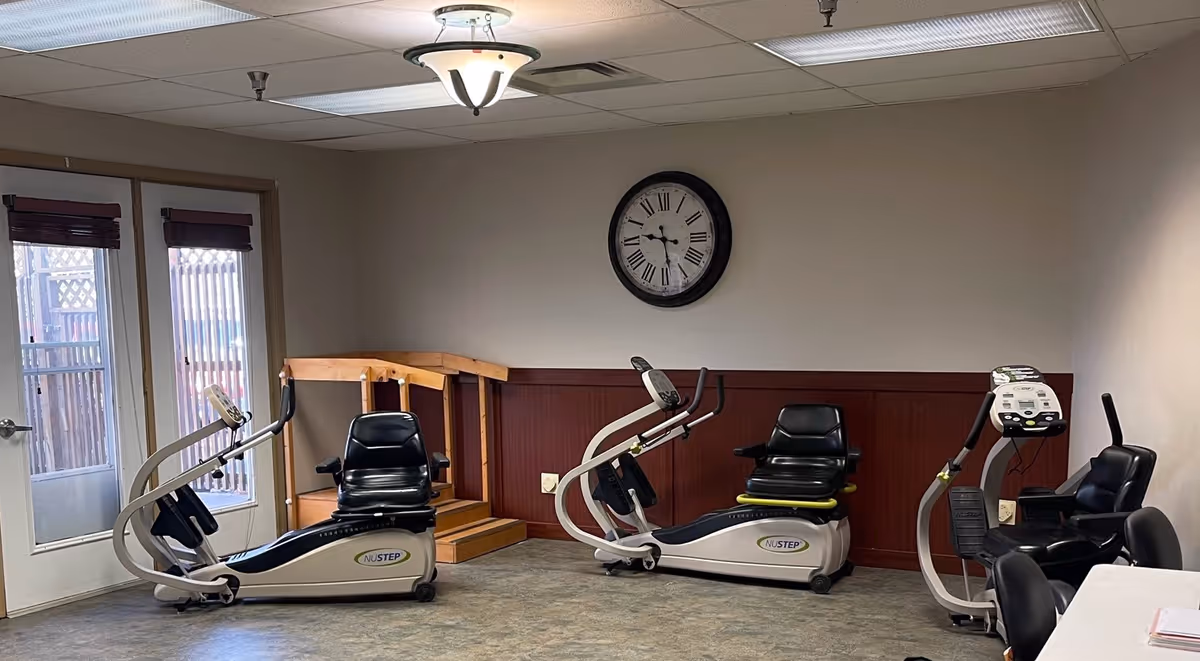 A small exercise room with three recumbent stationary bikes positioned near the walls. The room has a clock on the wall, a ceiling light fixture, and a door with windows leading outside. There is a wooden handrail and a small ramp in the corner.