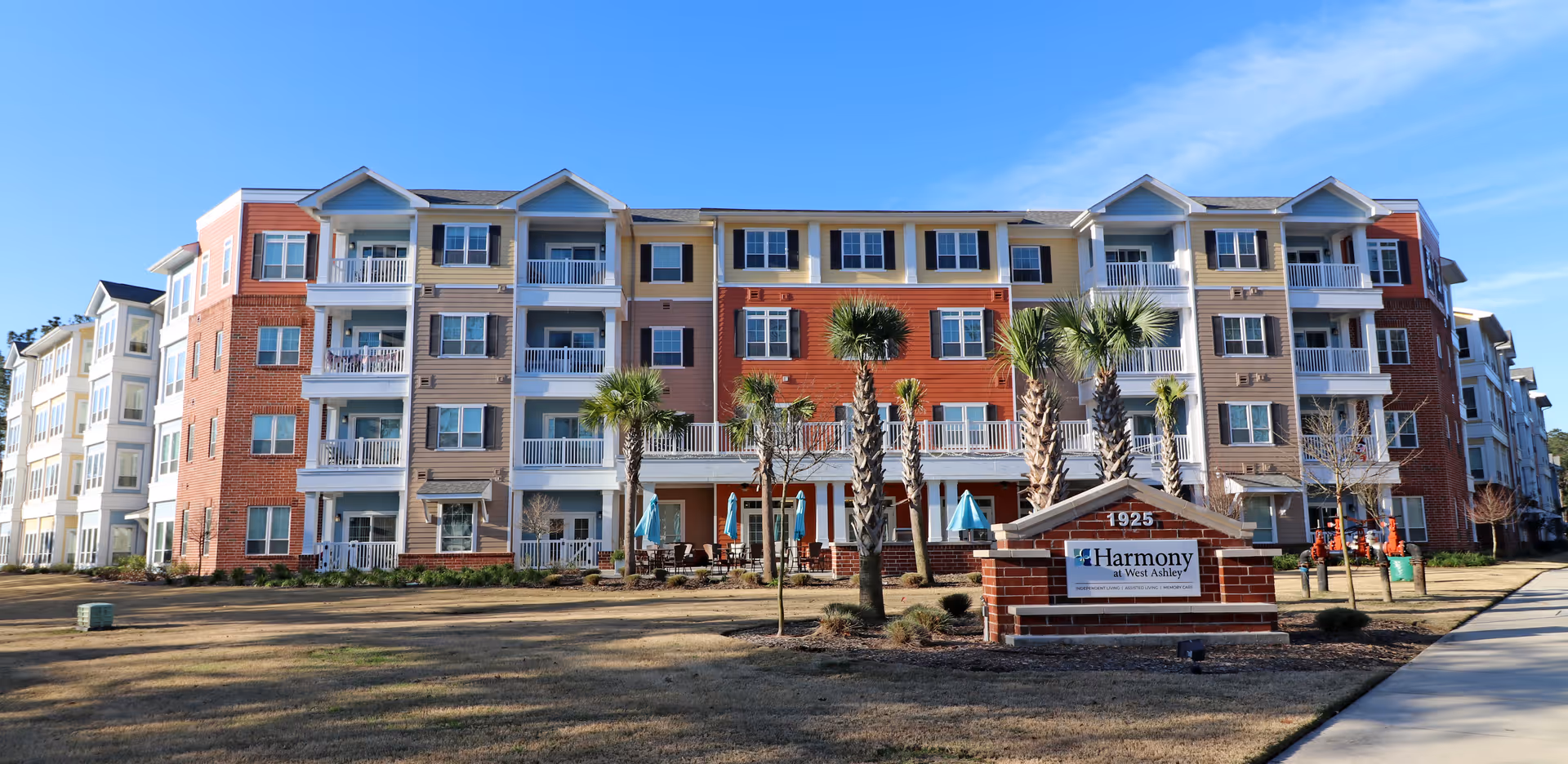 Exterior view of a multi-story senior living facility named Harmony at West Ashley with balconies, palm trees, and outdoor seating areas under blue sky.