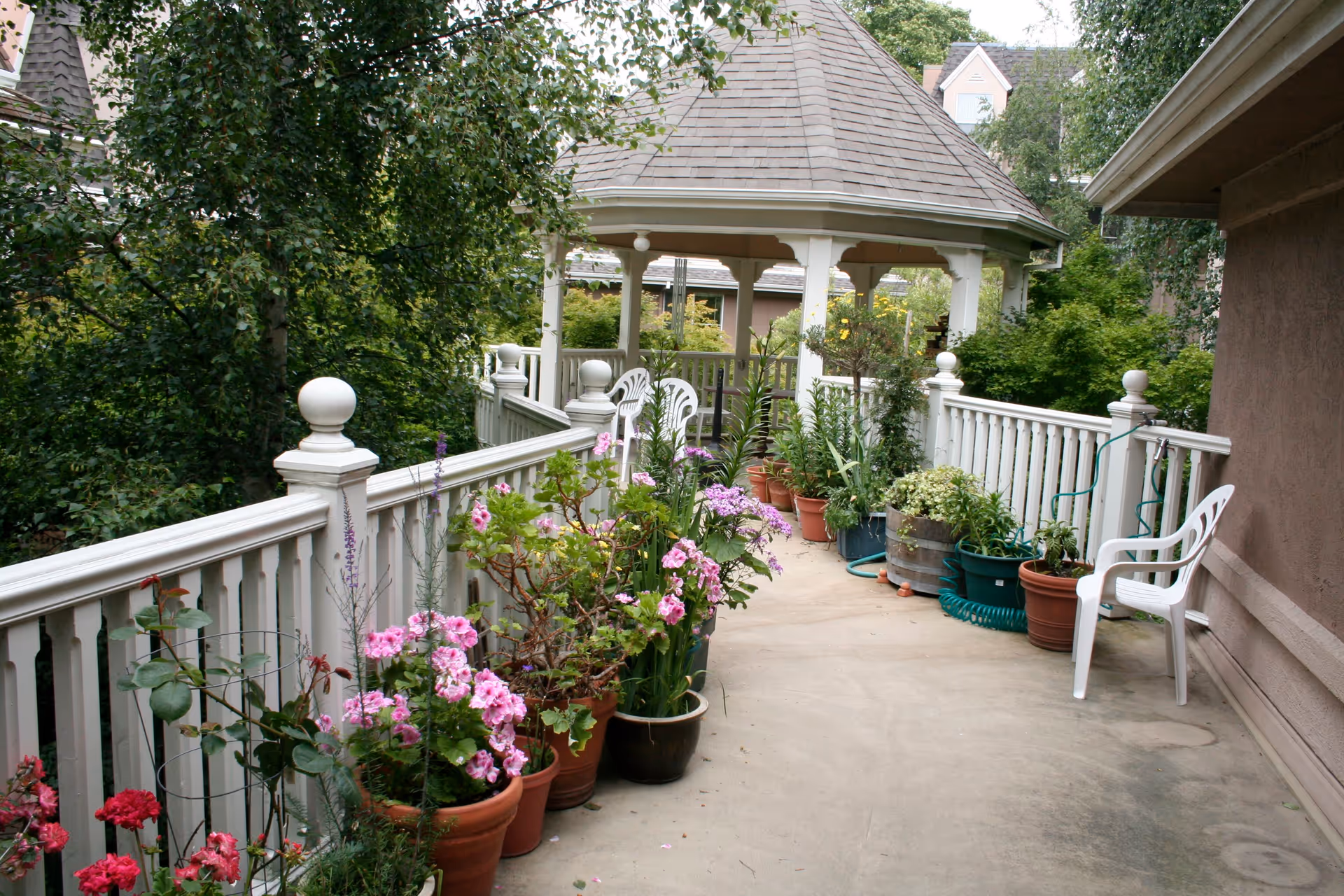 A garden walkway with a white railing lined with various potted plants and flowers leading to a gazebo. There is a white plastic chair on the right side near the building wall, and lush green trees and shrubs surround the area.