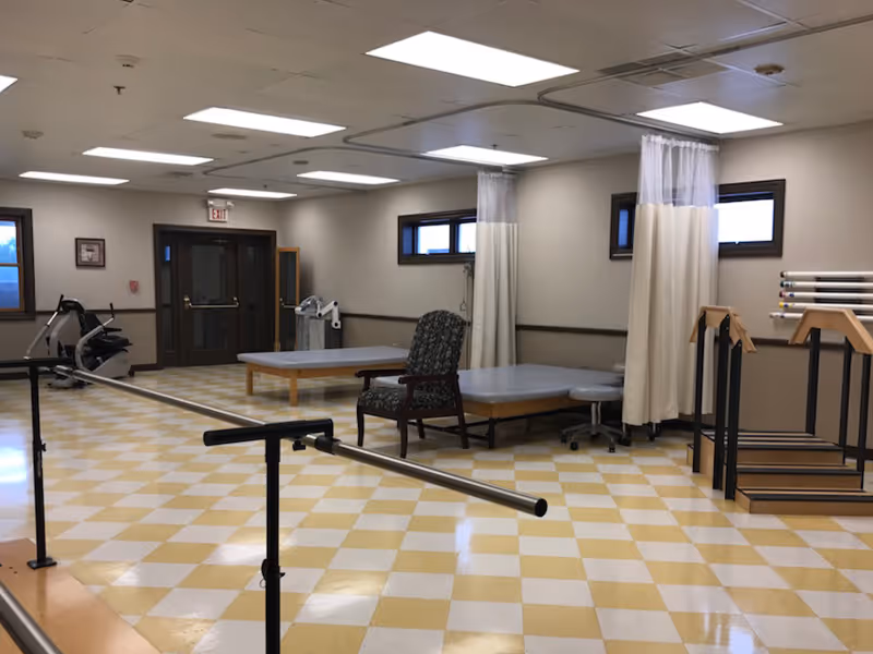 A rehabilitation therapy room with parallel bars, treatment tables, an exercise bike, and chairs under fluorescent lighting.