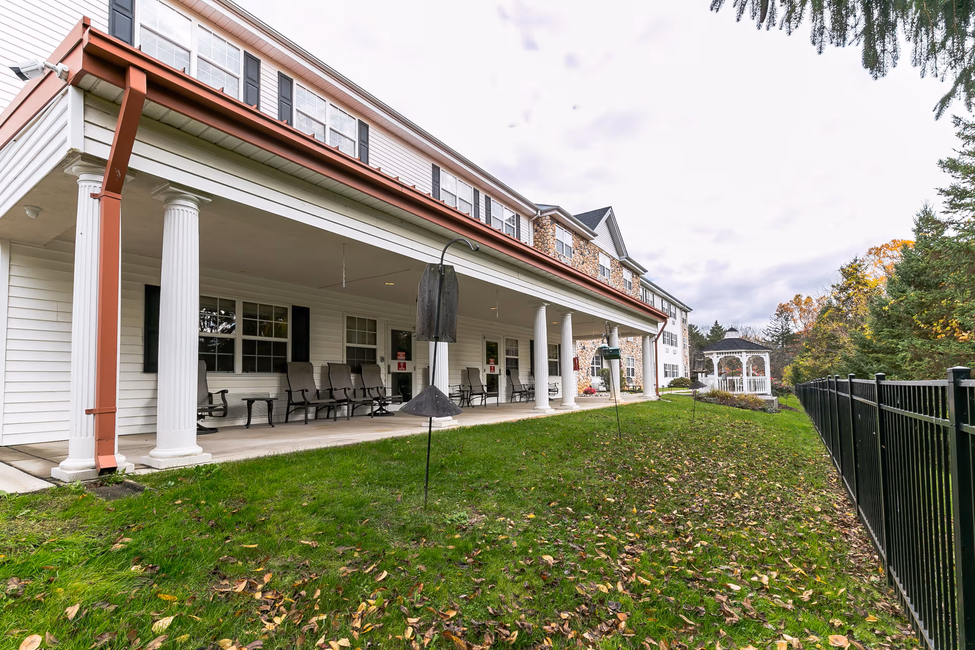 Exterior view of a senior living facility with a covered porch supported by white columns. Several rocking chairs are arranged on the porch. The building has white siding with black shutters and a stone accent wall. There is a grassy area with some fallen leaves in front of the porch, a black metal fence on the right, and a white gazebo further in the background surrounded by trees.