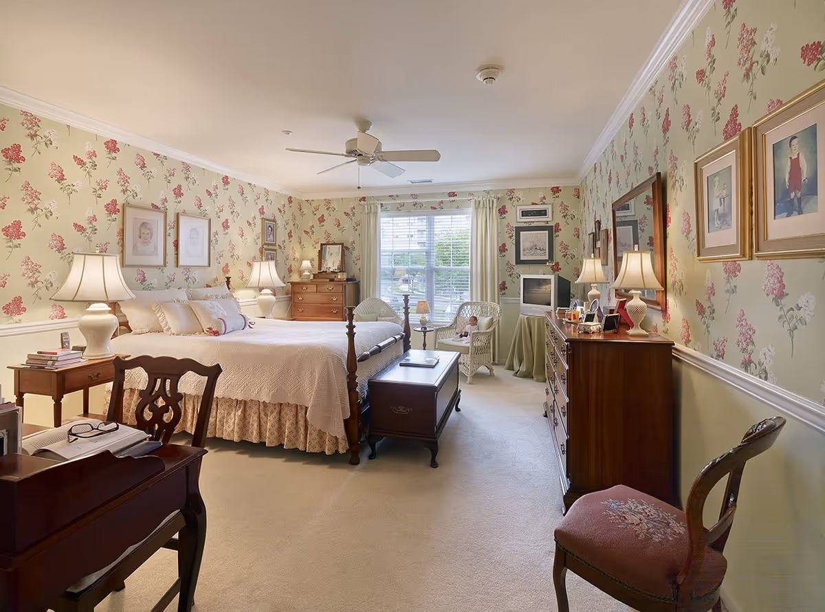 A cozy bedroom with floral wallpaper and a large window letting in natural light. The room features a wooden bed with white bedding, two bedside tables with lamps, a wooden dresser with a mirror, a small TV on a covered table, two wicker chairs with a small table between them, and a wooden desk with a chair. Several framed pictures hang on the walls.