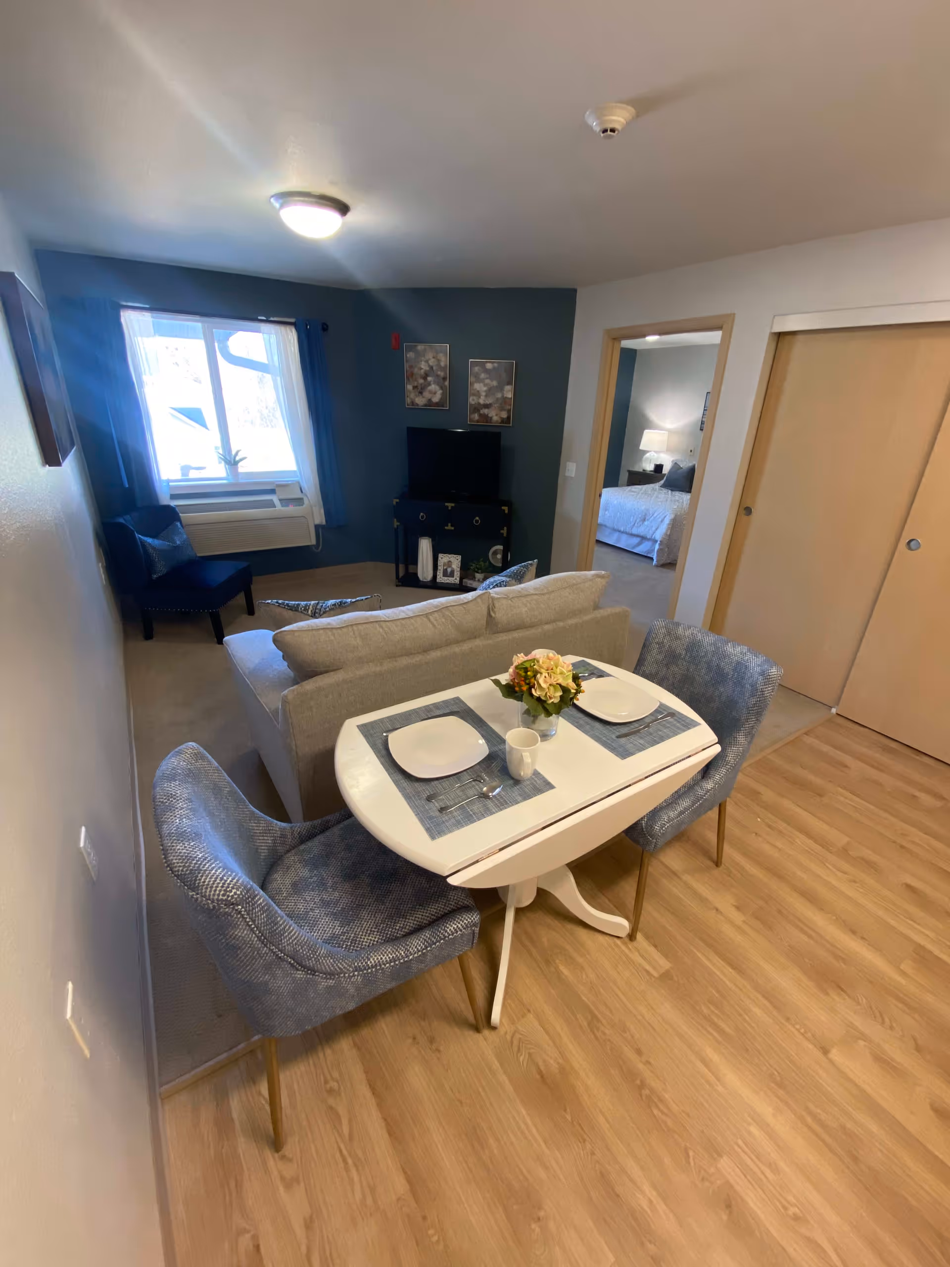 A cozy living and dining area in a senior living facility. The foreground features a small white dining table set for two with blue placemats, white plates, and a small flower arrangement. Two blue upholstered chairs are positioned at the table. Behind the table is a light gray sofa facing a TV on a dark blue stand against a blue accent wall. A window with blue curtains and an air conditioning unit is visible to the left. A doorway leads to a bedroom with a bed and nightstand with a lamp. Light wood flooring is in the dining area, and carpet is in the living and bedroom areas.
