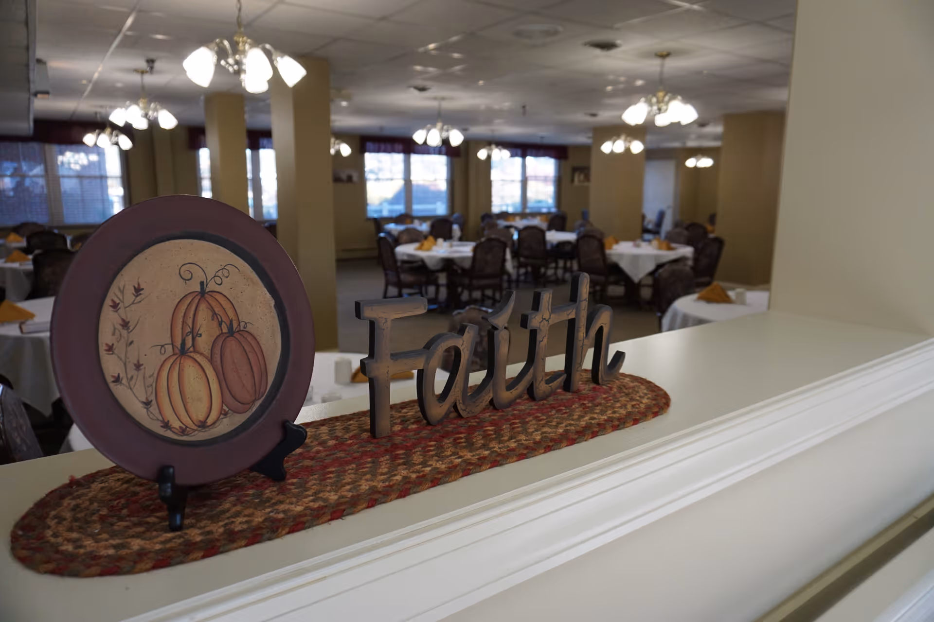 A dining room in a senior living facility with round tables covered in white tablecloths and set with napkins. In the foreground, there is a decorative plate with a pumpkin design and a wooden sign that says 'Faith' placed on a woven mat on a white ledge. The room is softly lit with chandeliers and has large windows letting in natural light.