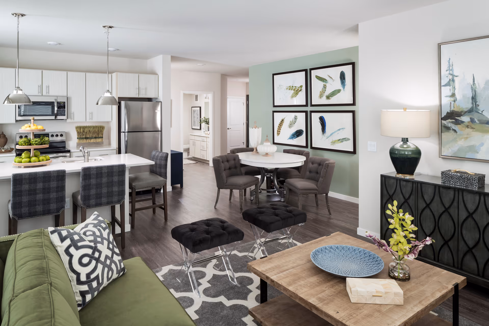 Open-concept living room and kitchen with a green sofa, breakfast bar with stools, round dining table, and decorative artwork.