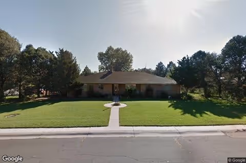 Single-story house with a manicured front lawn, central walkway, and trees on either side under a bright sky.