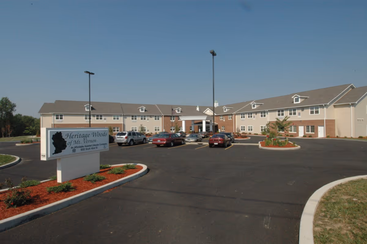 Exterior view of Heritage Woods of Mt. Vernon, showing a large two-story building with beige siding and brick accents. There is a parking lot with several cars in front of the building, landscaped areas with mulch and small shrubs, and a clear blue sky overhead. A sign in the foreground reads 'Heritage Woods of Mt. Vernon, An Affordable Assisted Lifestyle Community, 1033 South 42nd St.'