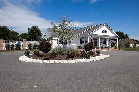 A single-story white building with a gray roof, featuring a covered entrance with white pillars and an American flag. In front of the building is a circular driveway with a landscaped island containing small bushes, plants, and a tree. The sky is partly cloudy and there are other houses and trees in the background.